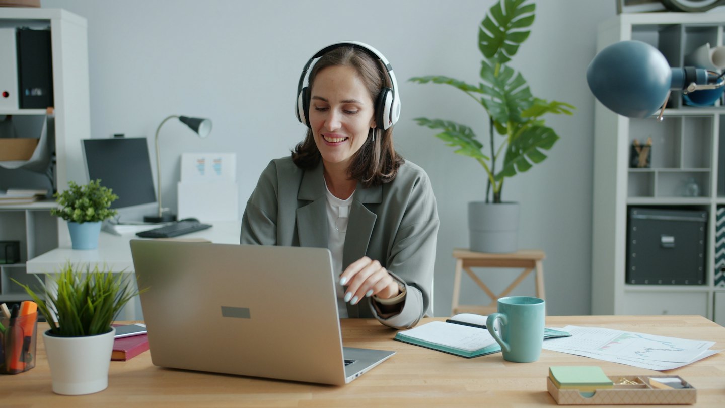 Woman with white headphones smiles while using a laptop at a desk.