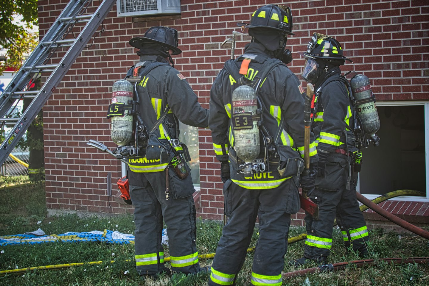 Three firefighters look at a brick building with a ladder.